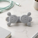 A gray silicone cord organizer with two smiling faces on a marble surface, surrounded by a light blue cable and a small