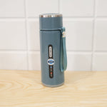 A blue insulated bottle with a silver cap and strap stands upright on a light wooden surface against a white tiled wall.