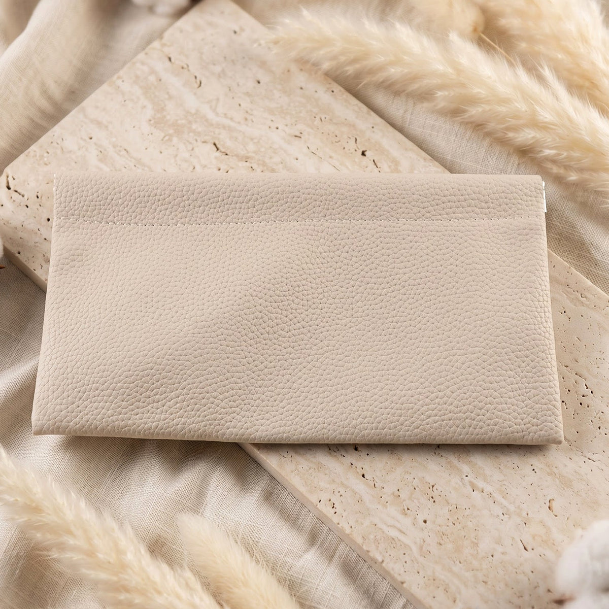 A cream textured faux leather pouch lying flat on a light-colored textured surface with white fluffy decorative elements.