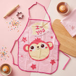 A pink monkey print apron is laid flat on a pink and white surface, surrounded by baking tools and cupcakes.