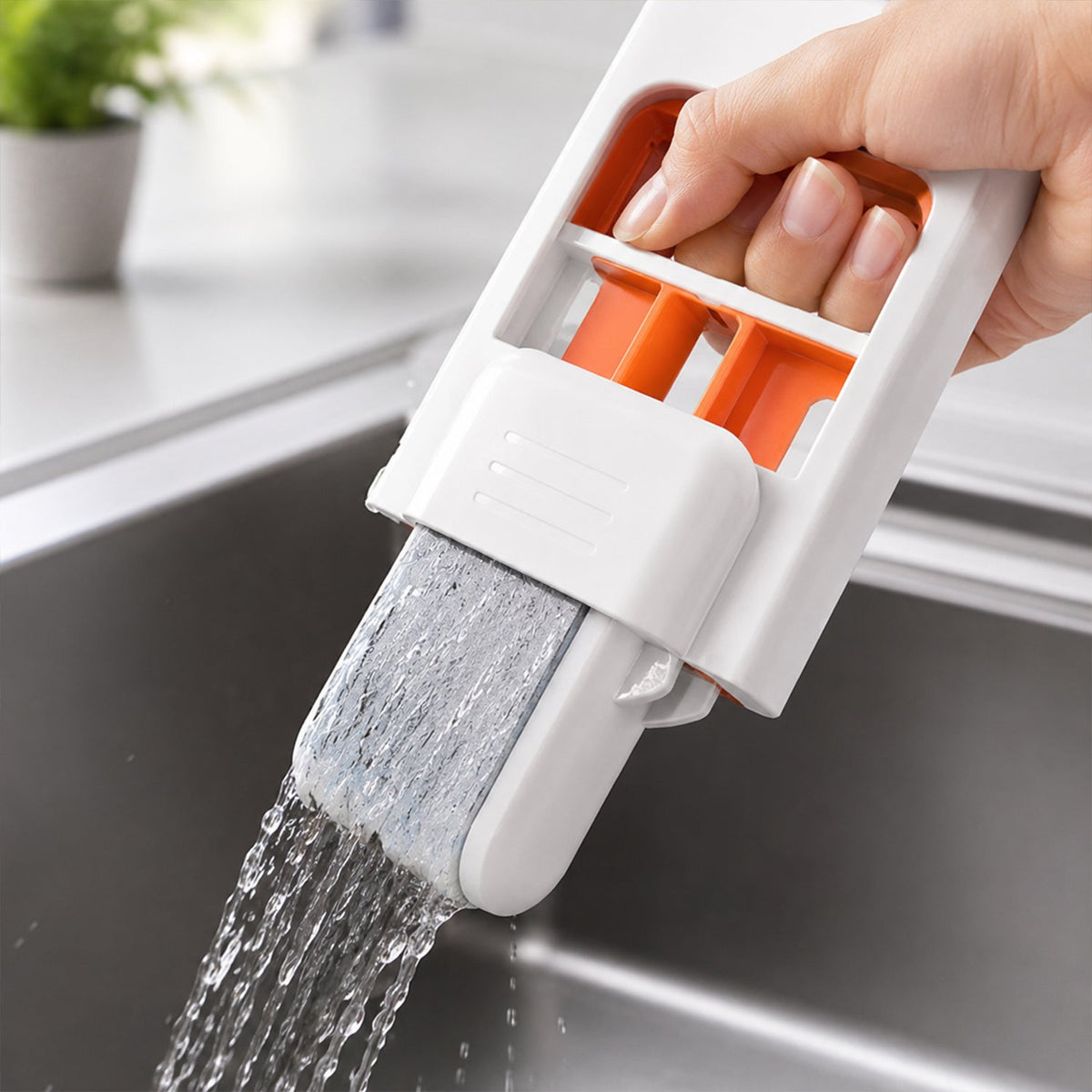 A hand uses the white and orange mini sponge mop to clean a window with suds, showing a blurred outdoor background.