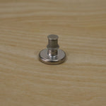 A silver metal button pin with its back lock attached, sitting on a light wooden surface.