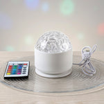 A white LED projection light with a clear textured dome and a remote control resting on a clear glass plate.