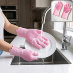 Hands wearing pink silicone gloves with scrubbing bristles washing a white plate under running water in a kitchen sink.