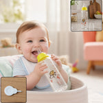 A happy baby in a high chair drinks from a clear glass baby bottle with a yellow nipple and monkey design.