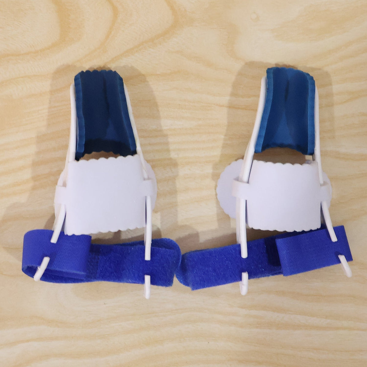 Two blue and white toe splints with blue straps are placed side by side on a light wooden surface.