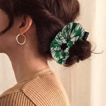 A person with brown hair wears a green and white leaf print scrunchie in a bun, with a gold hoop earring visible.