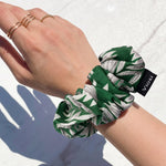 A person's arm wears a green and white leaf print scrunchie with a 'viosi' tag, along with gold rings on fingers, on a
