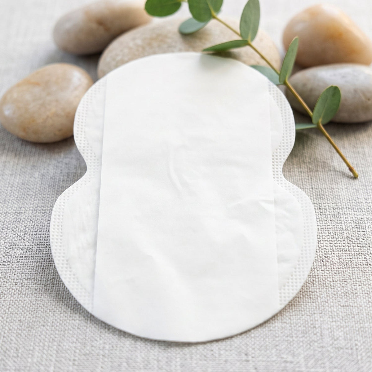 A single white, disposable underarm sweat pad with a smooth surface, surrounded by pebbles and a green sprig.