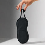A hand holds up a black, padded eye mask with an elastic strap against a grey and white background.