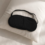 A black, padded eye mask with an elastic strap rests on a white pillow.