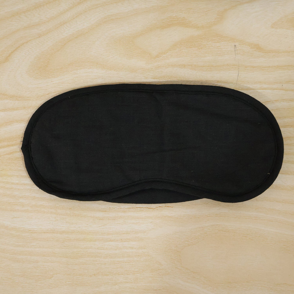 A black, padded eye mask with an elastic strap is laid flat on a light wooden surface.