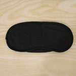 A black, padded eye mask with an elastic strap is laid flat on a light wooden surface.