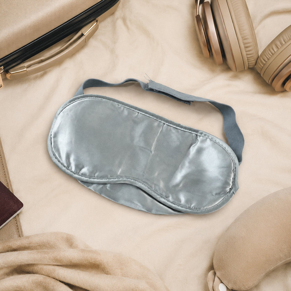 A grey satin sleeping eye mask with an elastic strap lies on a beige blanket next to headphones and a passport.
