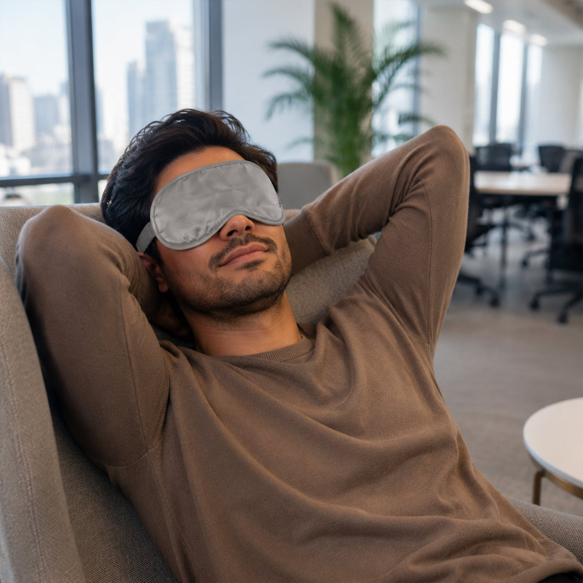 A man wearing a grey satin eye mask relaxes in a chair in a modern office with city views.