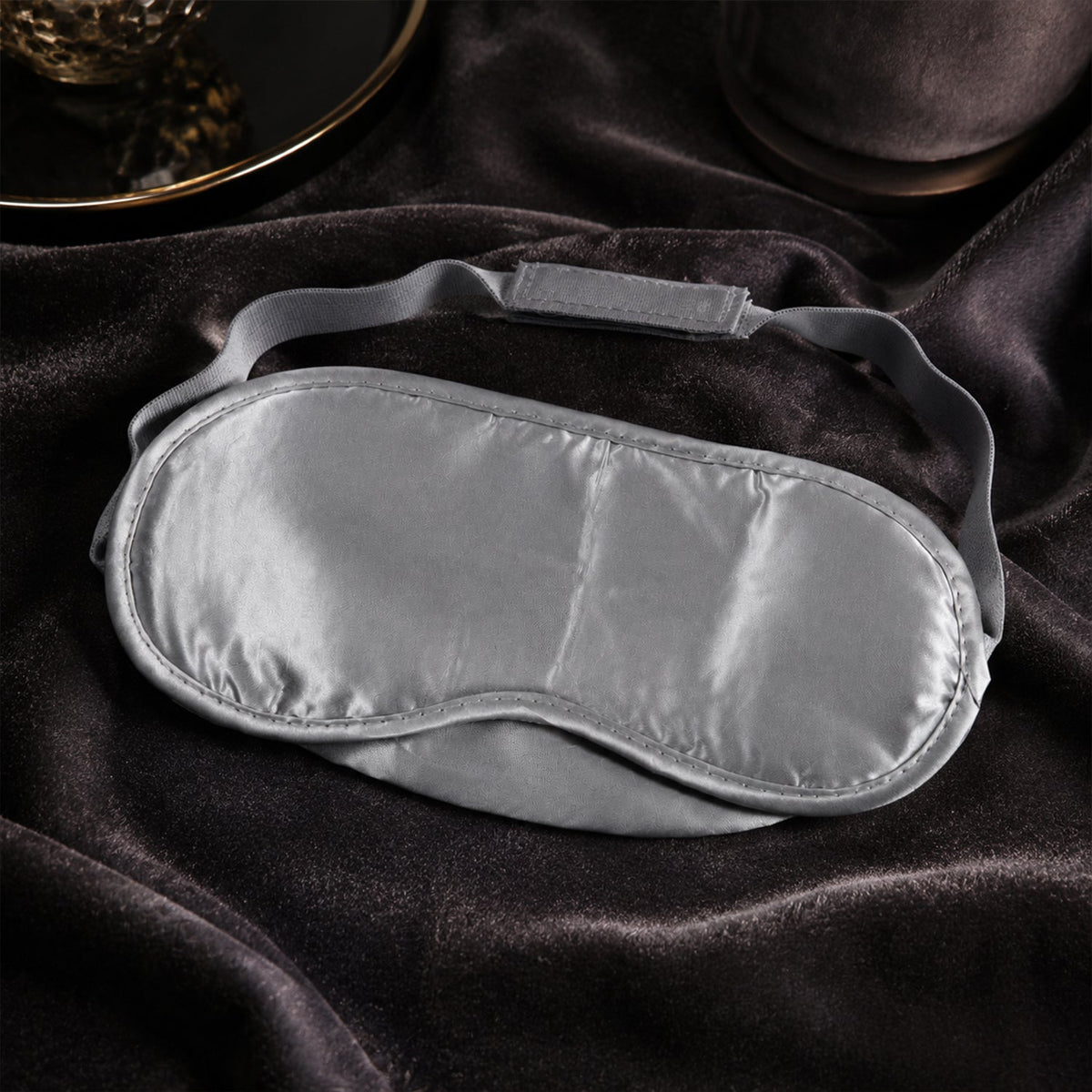 A grey satin sleeping eye mask with an adjustable elastic strap rests on a dark grey velvet surface.