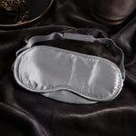 A grey satin sleeping eye mask with an adjustable elastic strap rests on a dark grey velvet surface.