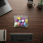 An overhead view of a clear plastic bag containing colourful thumbs-up clips on a wooden desk, surrounded by a laptop,