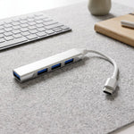A silver Type C to four-port USB hub with blue USB ports rests on a gray felt mat next to a keyboard.