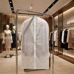 A translucent garment bag with a white zipper hangs on a metal rack in a clothing store with mannequins and racks of clothes.
