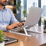 Man working on a silver laptop elevated by a white stand on a wooden desk in a modern office.