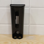 A black plastic bag dispenser sits on a wooden surface next to a clear adhesive hook on a white tiled wall.