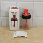 A glass honey dispenser with its box and a sheet of seasoning labels on a light wooden surface.