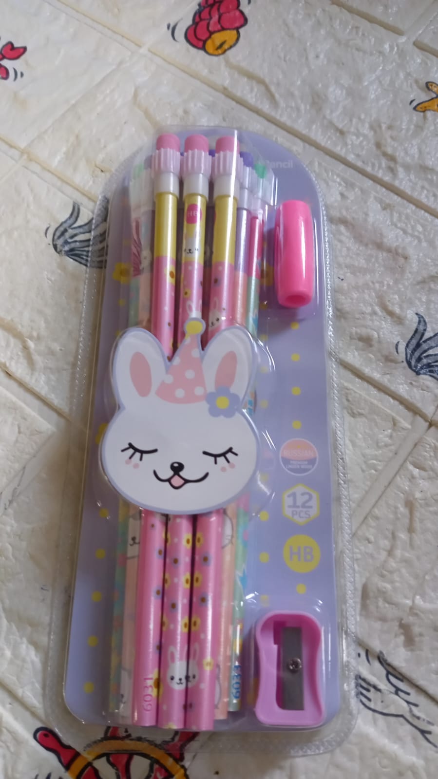 A clear plastic package with a bunny face holds twelve pastel pencils, a pink eraser, and a pink sharpener, on a textured