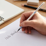 A hand writes "Hello World" on white paper with a white and blue pen refill, with a notebook and eraser in background.