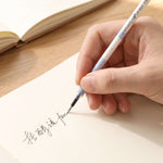 A hand writes in a foreign language on a light-colored paper with a white and blue pen refill, with a notebook.