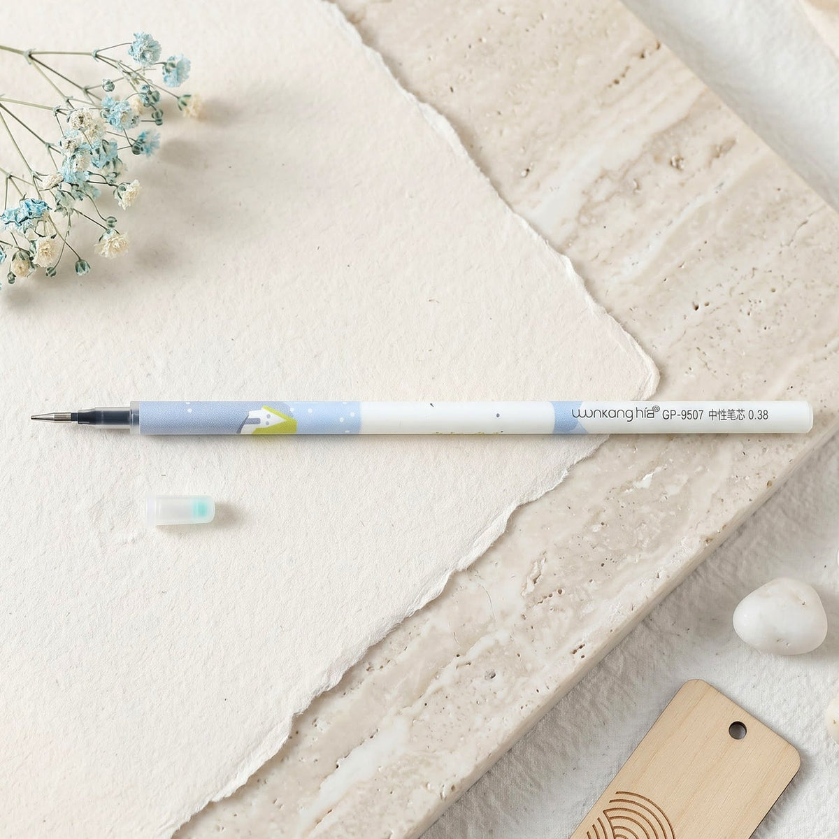 A white and blue pen refill with a light blue cap on textured white paper, with small blue flowers and a white stone.