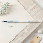 A white and blue pen refill with a light blue cap on textured white paper, with small blue flowers and a white stone.