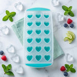 A clear and teal heart-shaped ice cube tray rests on a marble surface, surrounded by loose heart ice, mint leaves,