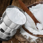 The grinder's stainless steel bowl is filled with white flour, next to a wooden bowl of flour and a spoon.