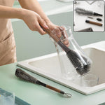 A person cleans a clear glass carafe with a long-handled grey brush, while a second brush rests on the counter.