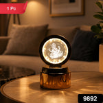A golden lamp with a glowing Ganesha crystal sits on a round wooden table, with a blurred living room in the background.