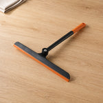 An orange and black squeegee with a pivoting head rests on a light wood surface next to a folded white towel.