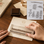 Hands press a clear stamp with a floral border design onto an elegant cream-colored invitation card, with an ink pad nearby.