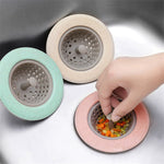 Three silicone sink strainers in mint green, beige, and pink, with a hand removing food scraps from the pink one.