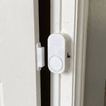 A white magnetic door alarm is installed on a slightly ajar white door frame, with the two parts separated.