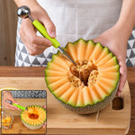 A person uses a green-handled dual-ended tool to scoop seeds from a carved cantaloupe melon on a wooden cutting board.