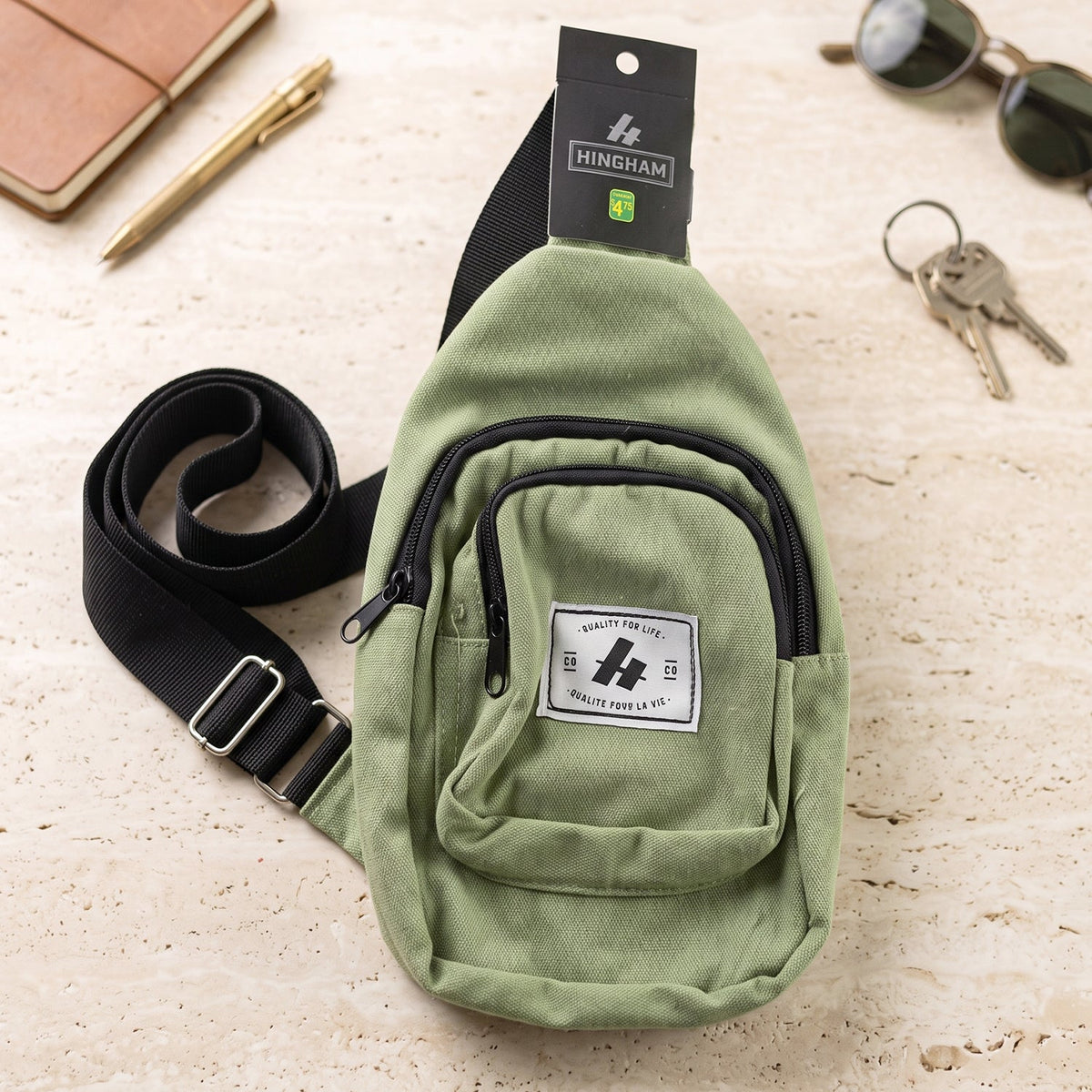 An olive green canvas sling bag with a black strap, Hingham tag, sunglasses, keys, and a pen on a speckled surface.