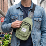 A man in a denim jacket wears an olive green canvas sling bag with a black strap and a Hingham tag on the shoulder.