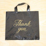 A black plastic 'Thank You' bag with gold script is displayed flat on a light-colored wooden background.