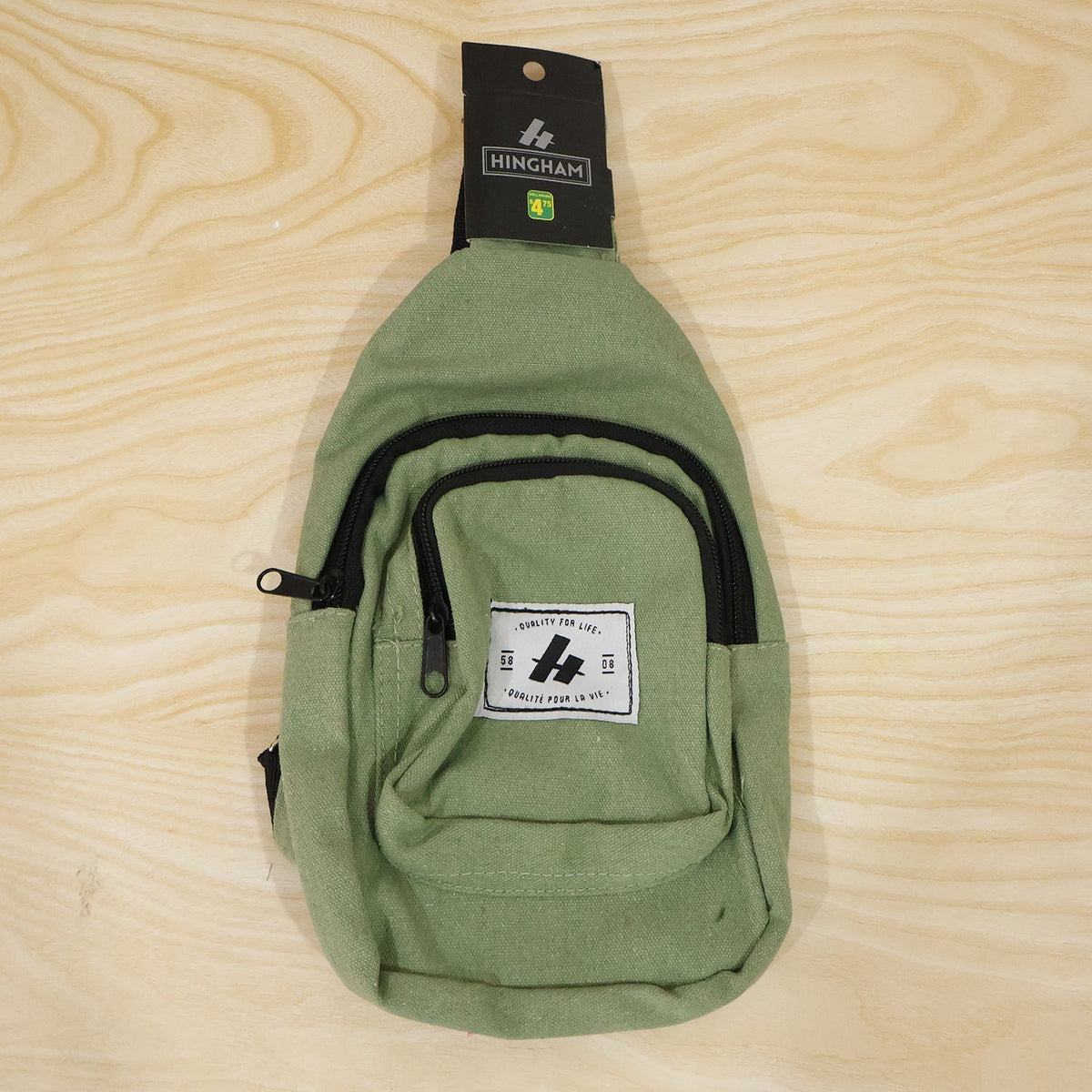 An olive green canvas sling bag with a front pocket and black zippers, displayed on a light wooden surface.