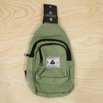 An olive green canvas sling bag with a front pocket and black zippers, displayed on a light wooden surface.