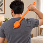 A person uses an orange silicone back scrubber with a long handle on their back in a bedroom setting.