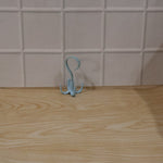 A light blue rotating multi-hook hanger sits on a light wooden surface against a white tiled wall.