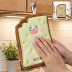 A person's hands are shown drying on a cartoon cheese toast-shaped microfiber towel hanging from a kitchen cabinet knob