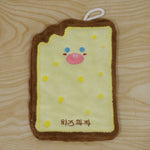 A cartoon cheese toast-shaped microfiber towel with a brown crust and yellow center, featuring a face, lies on a wooden